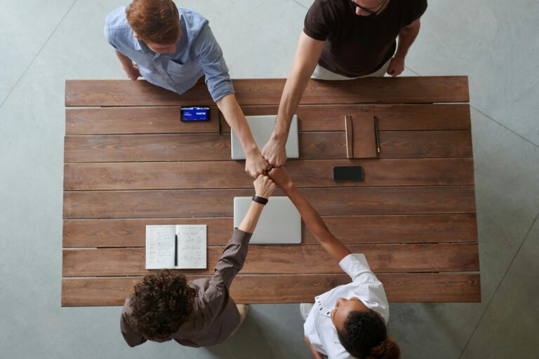Quatro pessoas unem os punhos sobre uma mesa de trabalho, vistas de cima
