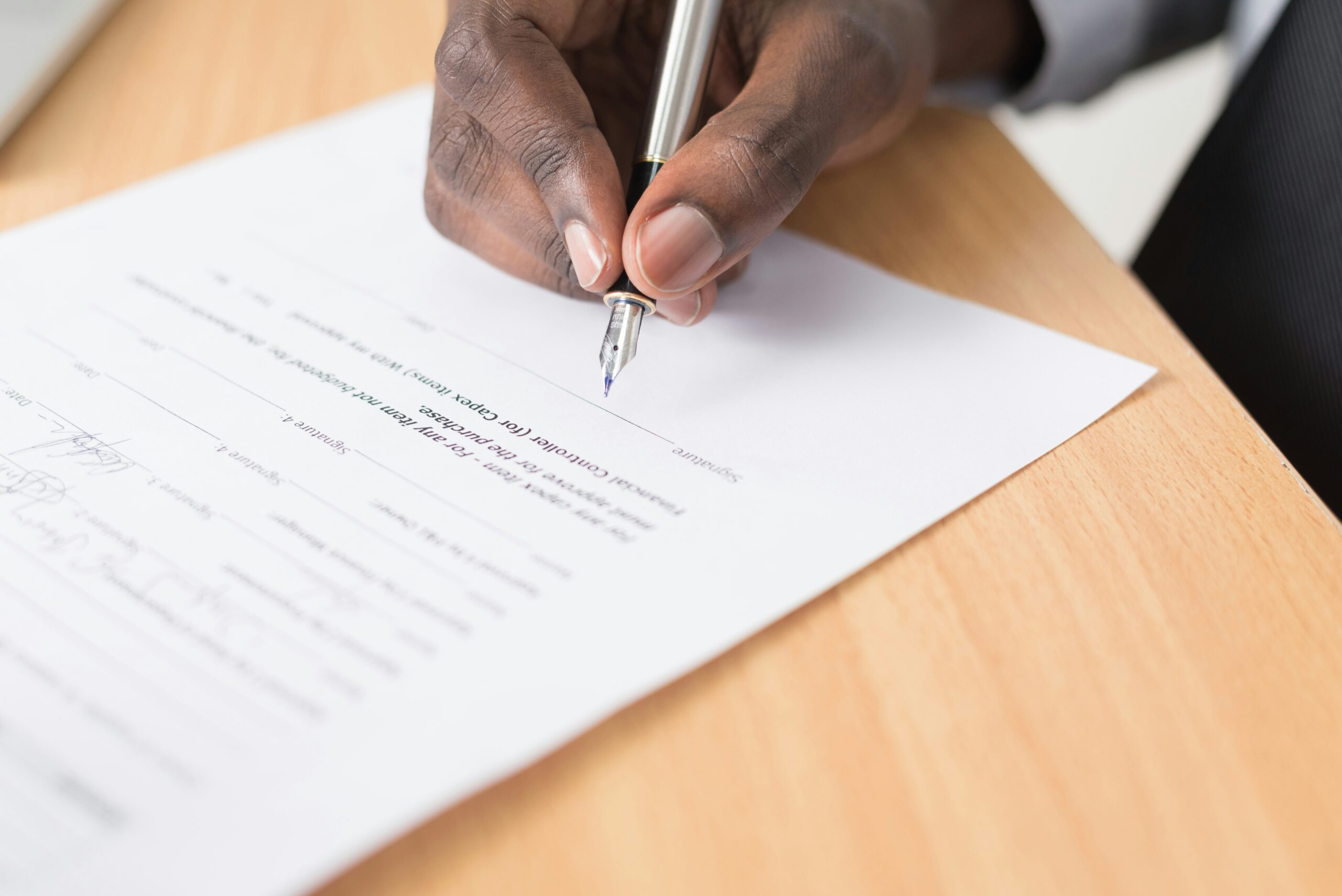 Homem negro com terno cinza assinando documento sobre mesa de madeira clara com caneta-tinteiro para formalizar cláusula de não concorrência empresarial.